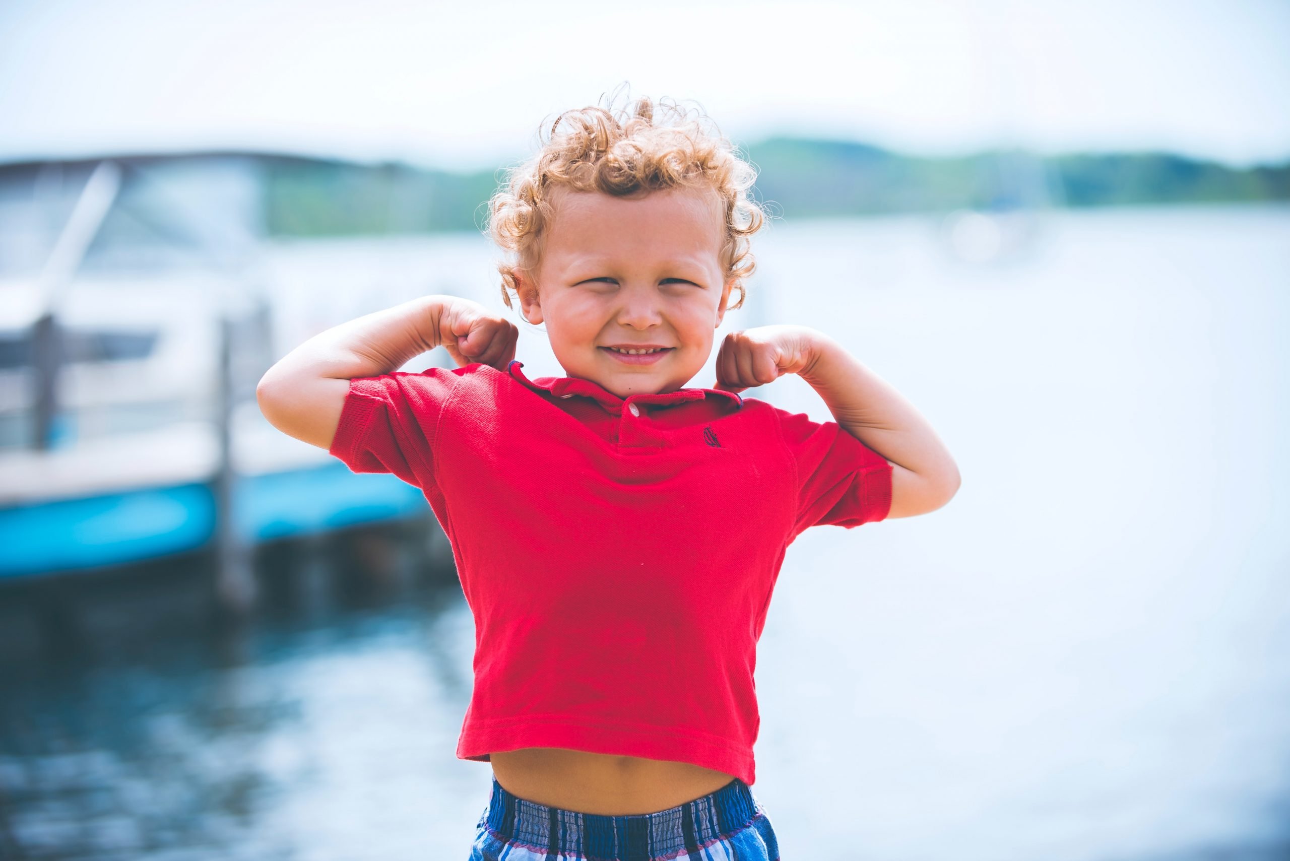 little boy in red t-shirt and curly hair flexing his arm muscles for the camera 