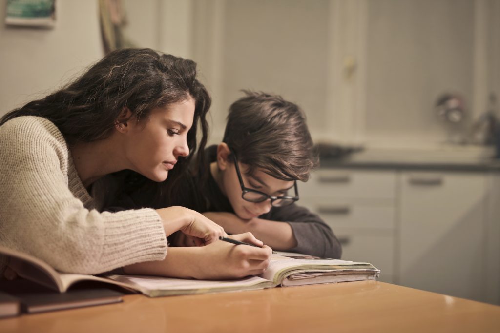 woman leaning over to point at the notebook a boy is holding with a pencil