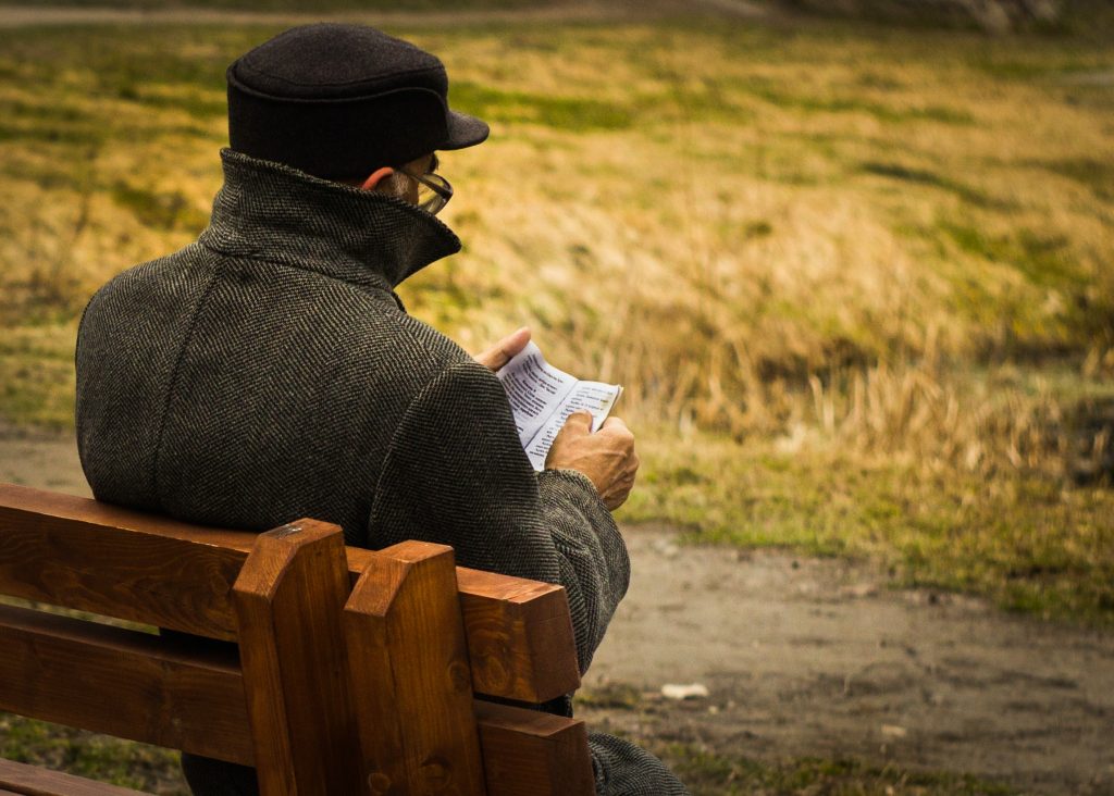 man with a hat and coat siting on a bench reding a small book 