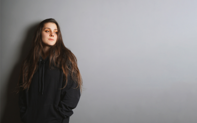 girl with brown hair wearing black hoodie standing against the wall