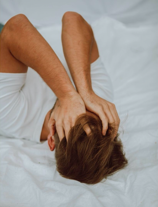 man in white tshirt holding his face with hands while lying on bed