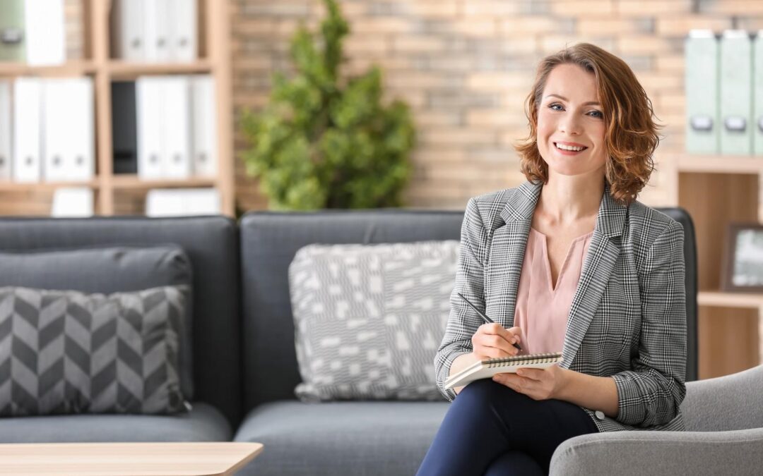 woman with short hair wearing a striped blazer smiles while sitting on a couch