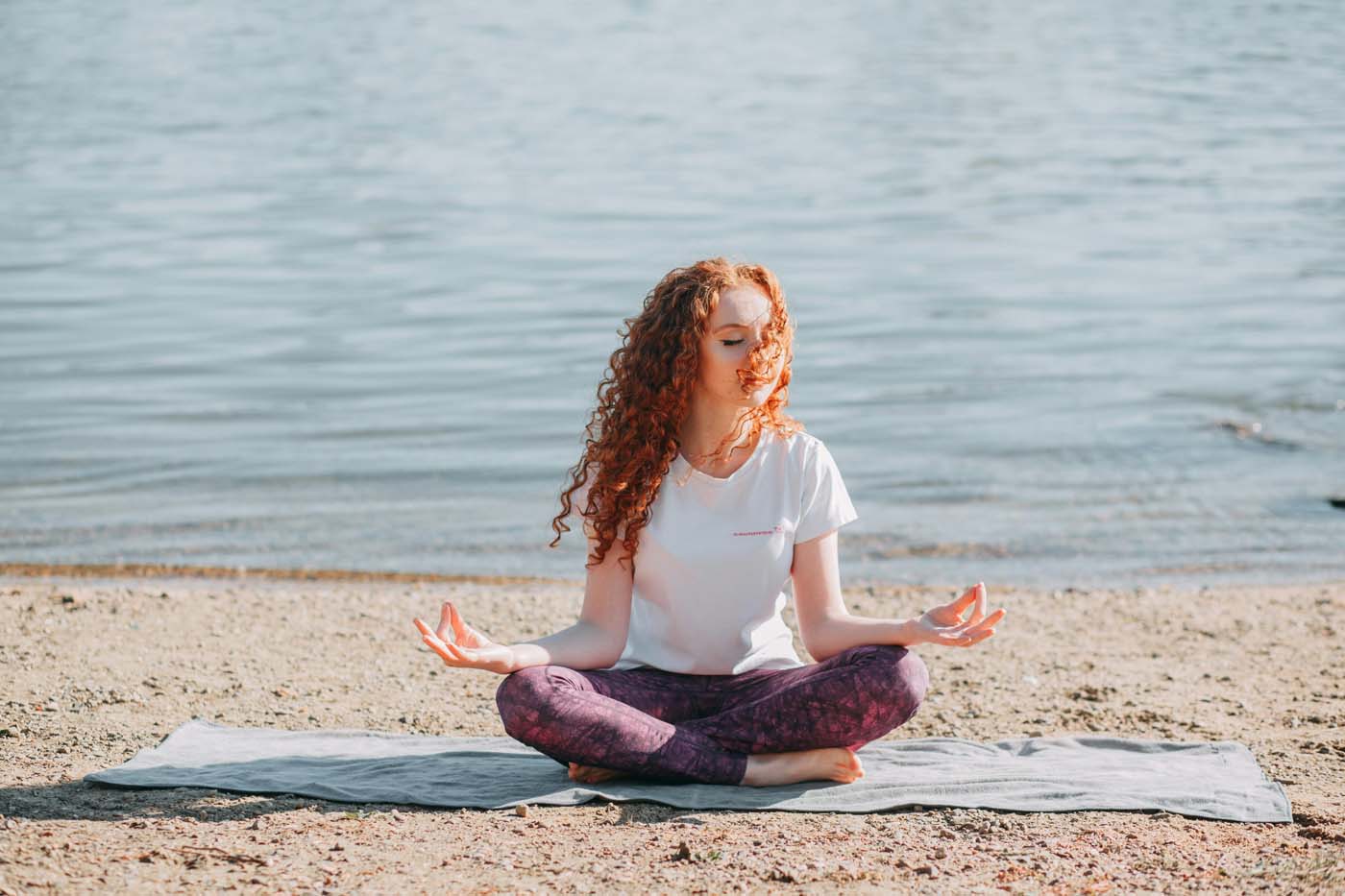 Image of a peaceful young woman practicing meditation while sitting cross legged on a yoga mat near water. Learn to embrace your spirituality and your higher self with the help of a skilled spiritual therapist in Simi Valley, CA.