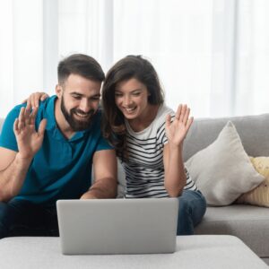 A cheerful man and woman sitting on a sofa, waving at a laptop screen