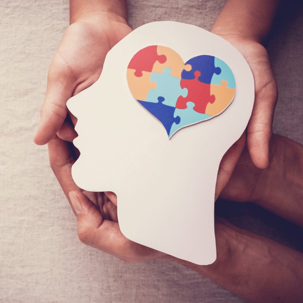 Hands holding a paper cutout of a human head in profile, with a heart-shaped brain made of colorful puzzle pieces