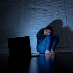 A person sitting on the floor in a dark room, illuminated by a laptop screen, appears distressed