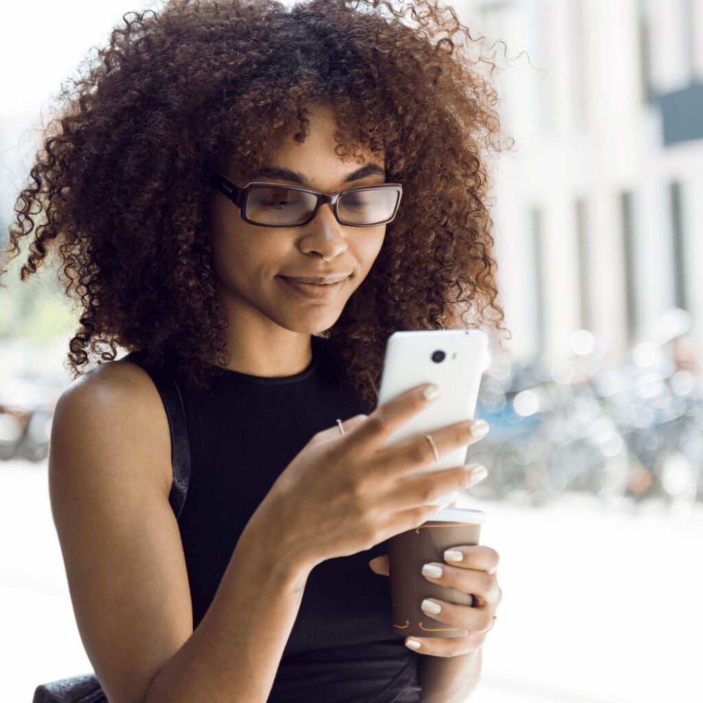 A woman with curly hair and glasses holds a to-go cup and looks at her smartphone while standing outside.