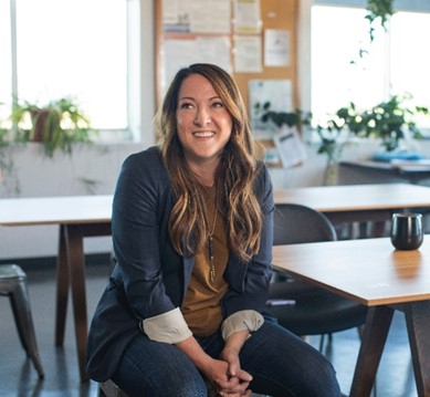 A woman with long hair sits on a chair in a casual office setting.