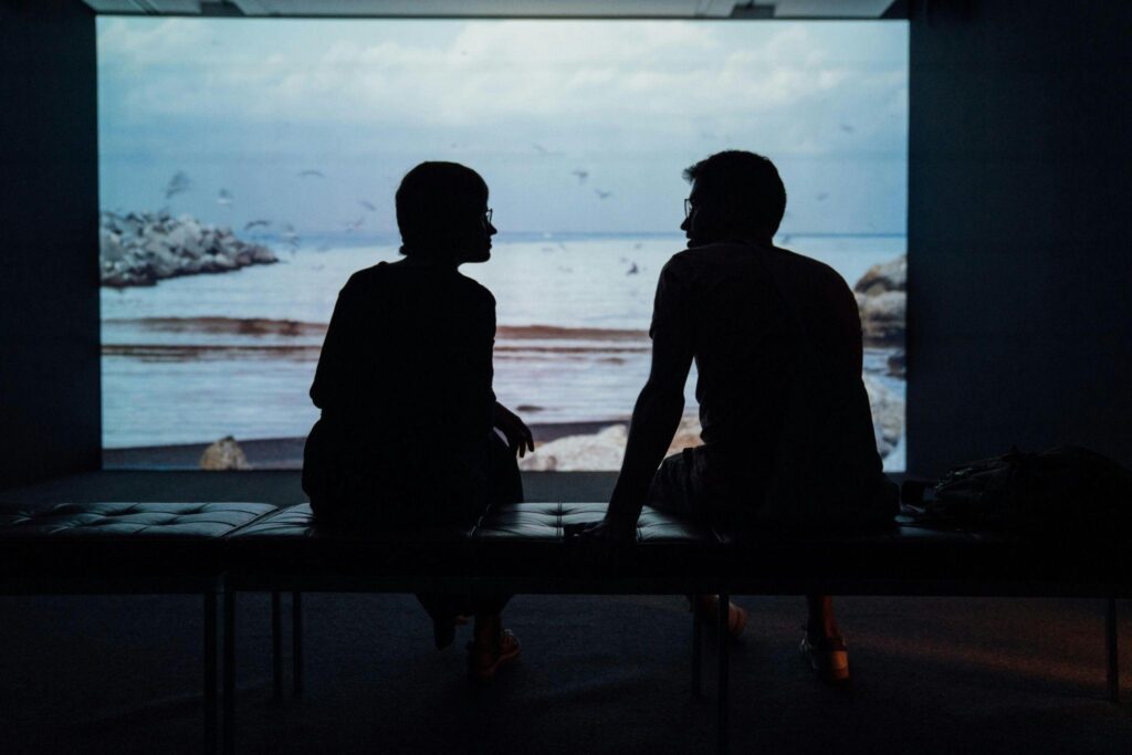 Two people sit on a bench in a dimly lit room, silhouetted against a large screen displaying an ocean scene.