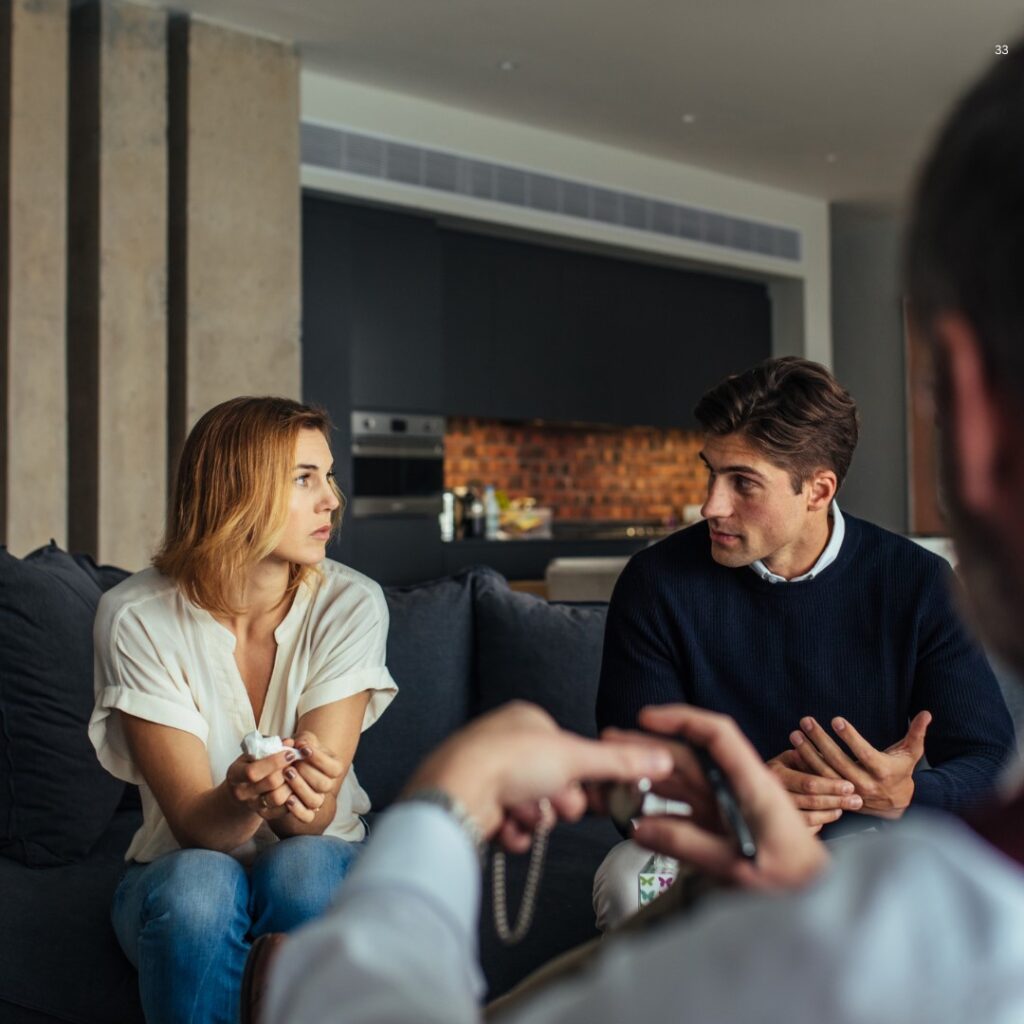 A woman and a man are sitting on a sofa having a serious conversation. Another person in the foreground is taking notes.