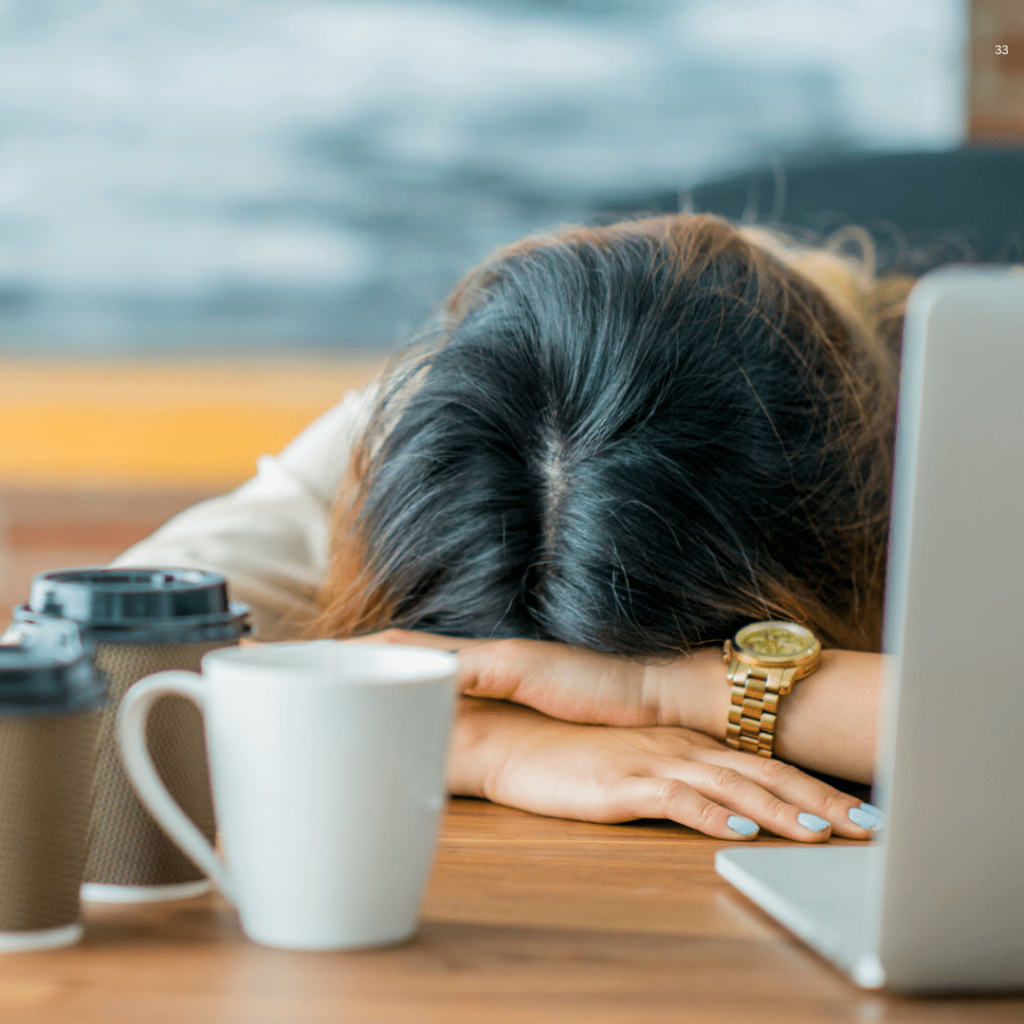 A woman rests her head on a desk, sleeping peacefully beside an open laptop, surrounded by a quiet workspace.