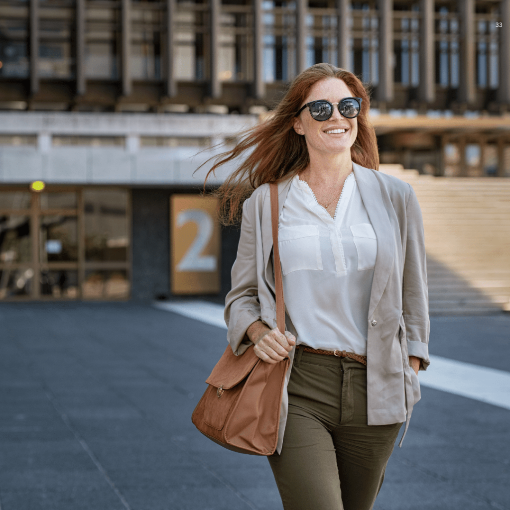 A woman wearing sunglasses and a blazer confidently walks down the street, exuding style and sophistication.