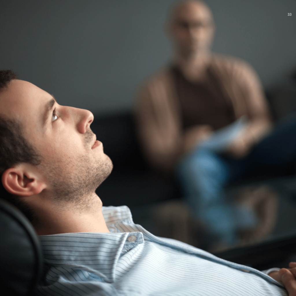 Person in a striped shirt reclining on a couch, with a blurred figure in the background suggesting a therapy or counseling session.