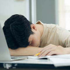 A man rests his head on a table, sleeping peacefully at his desk, surrounded by papers and a computer.