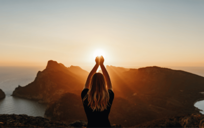 A woman stands triumphantly on a mountain peak at sunset, arms raised in celebration against a vibrant sky.