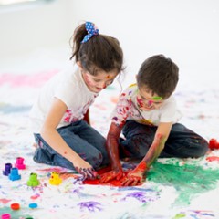 Two children sitting on the floor, engaged in finger painting. They are covered in colorful paint, with various paint pots scattered around them.