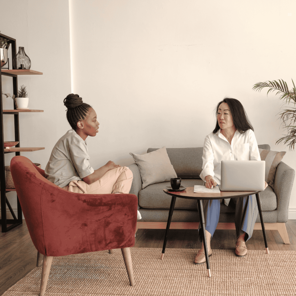 Two people in a modern living room, one sitting on a sofa with a laptop, the other on a red chair.