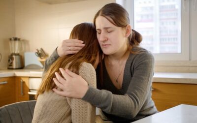 Two people sit in a kitchen, with one comforting the other by embracing them in a supportive hug.