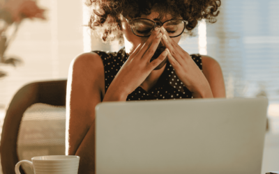 woman with curly hair and glasses, sitting at a desk, pinching the bridge of her nose in frustration or stress.